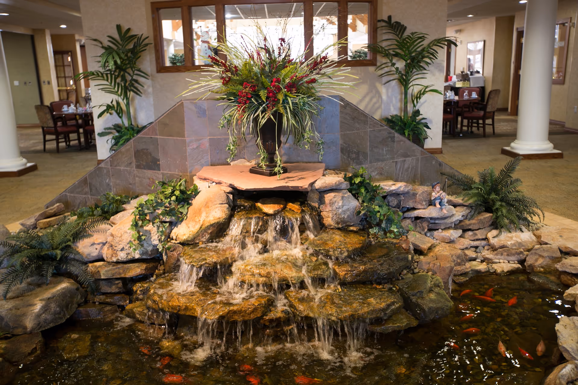 Indoor water feature with cascading waterfall over rocks into a pond with orange fish, surrounded by plants and greenery. In the background, there are tables and chairs in a common area with beige walls and carpeted floors.