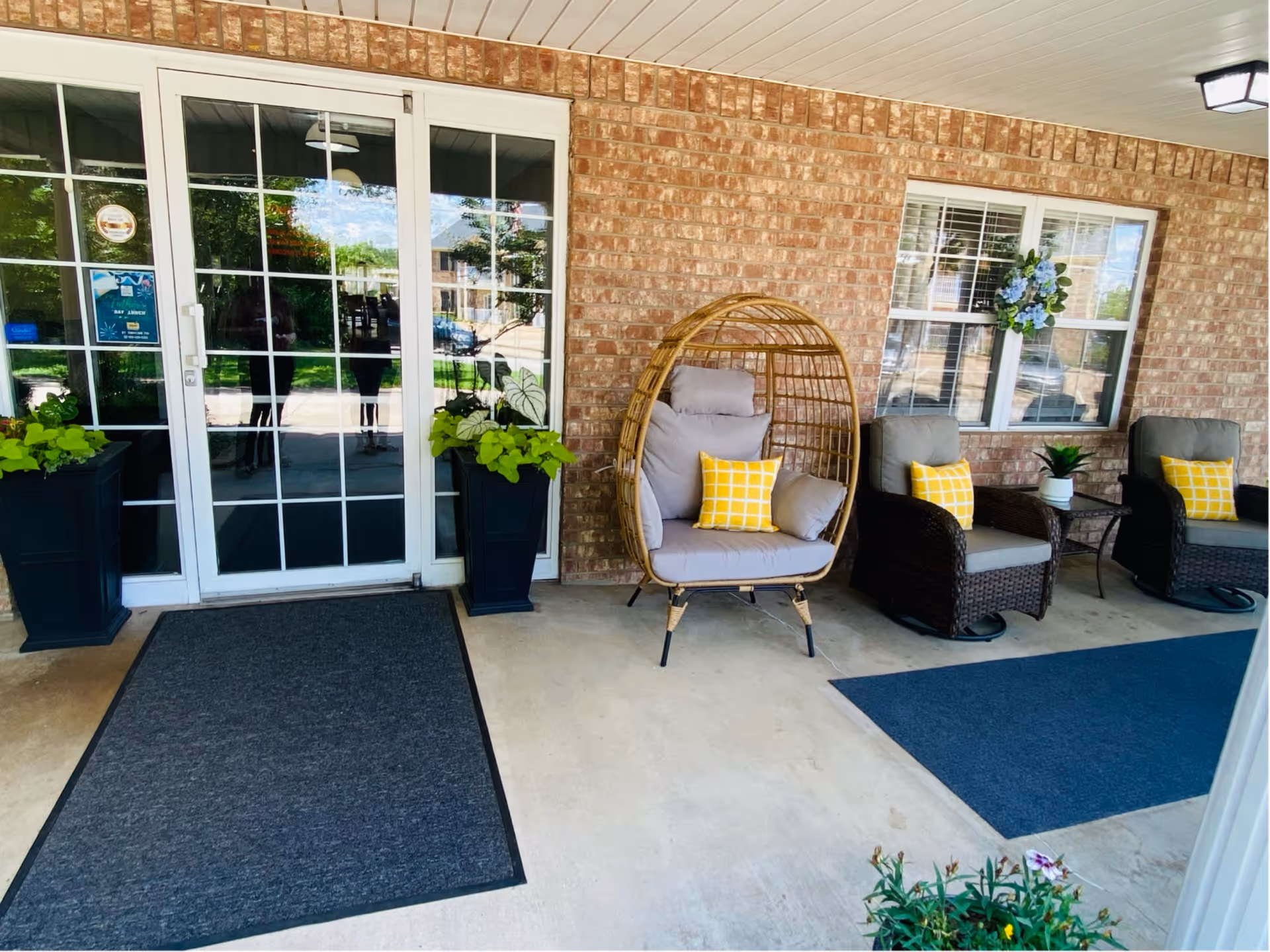 Covered brick entrance at Hopkins Court with glass double doors, potted plants, and wicker seating including an egg-shaped chair with yellow pillows.