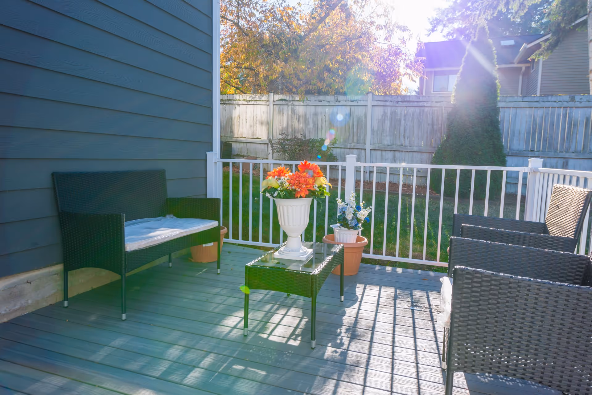 Sunlit backyard deck with wicker patio chairs, a small table and potted flowers against a white railing and wooden fence.