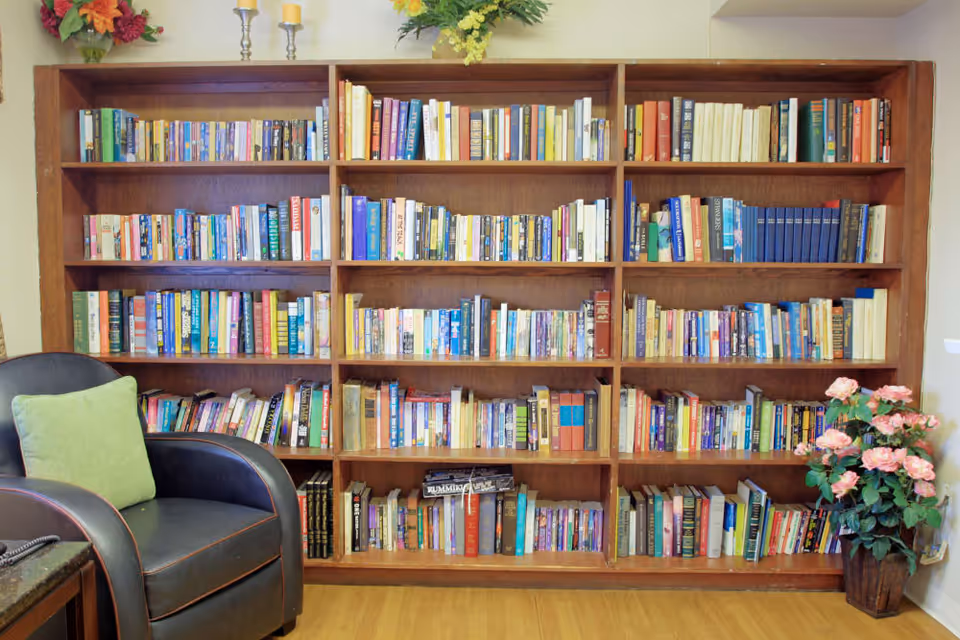 A large wooden bookshelf filled with numerous books of various sizes and colors. To the left of the bookshelf is a black armchair with a light green cushion. On the right side of the bookshelf, there is a pot with pink flowers. The floor is wooden, and there are decorative flowers and candles on top of the bookshelf.