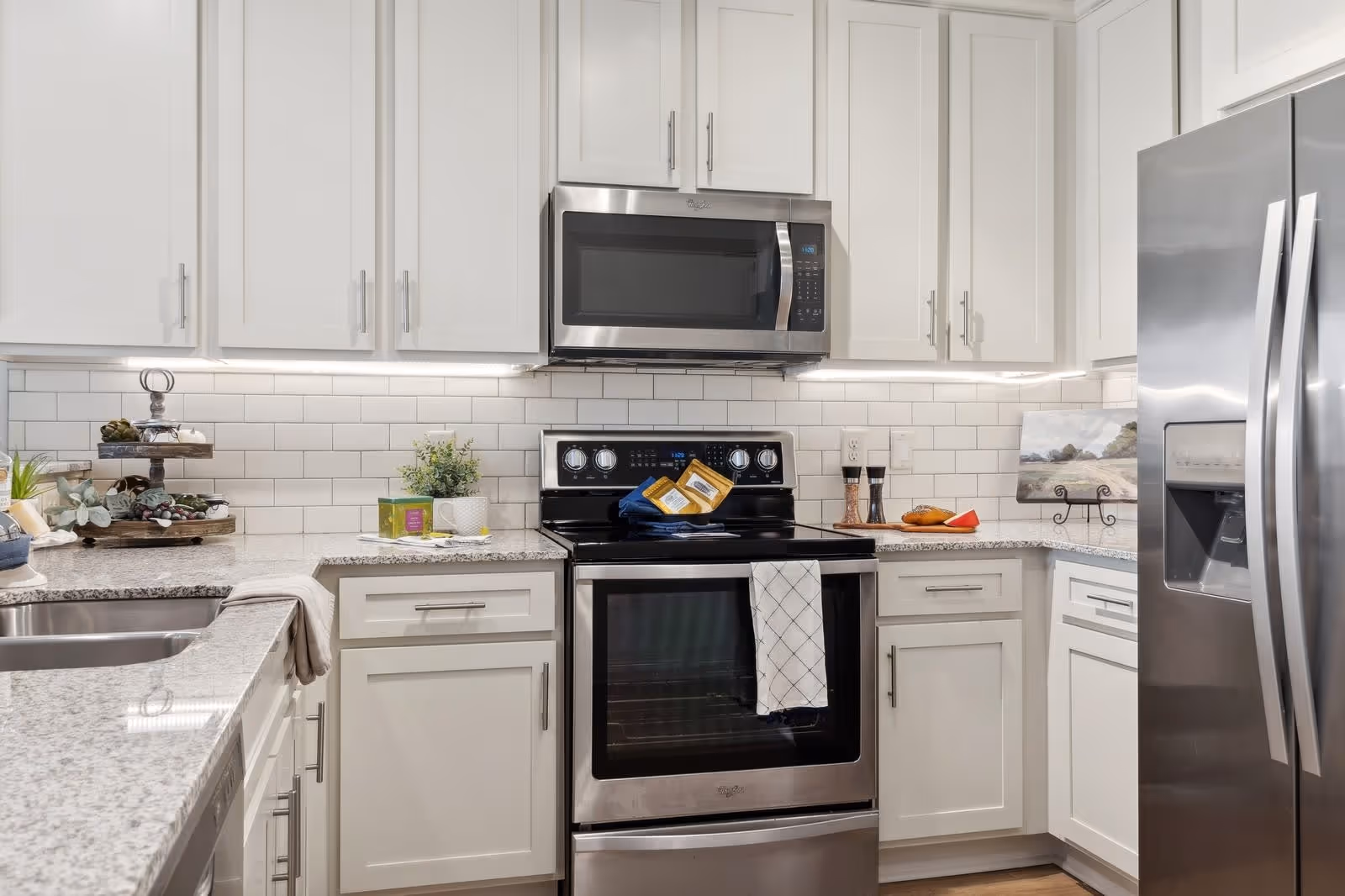 Modern kitchen with white cabinets, granite countertops, stainless steel microwave, oven, and refrigerator. The backsplash is white subway tile. There are kitchen towels hanging on the oven handle and various decorative items on the countertops.