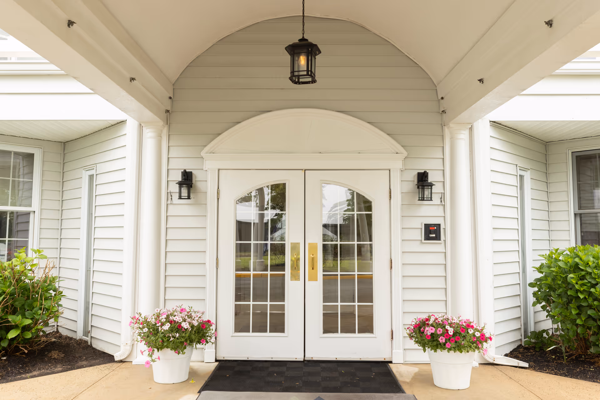 Entrance to a building with white double doors featuring glass panes, flanked by two white columns and two potted plants with pink flowers. The exterior walls are light gray with white trim, and there are two black wall-mounted lanterns on either side of the doors and a hanging lantern above.