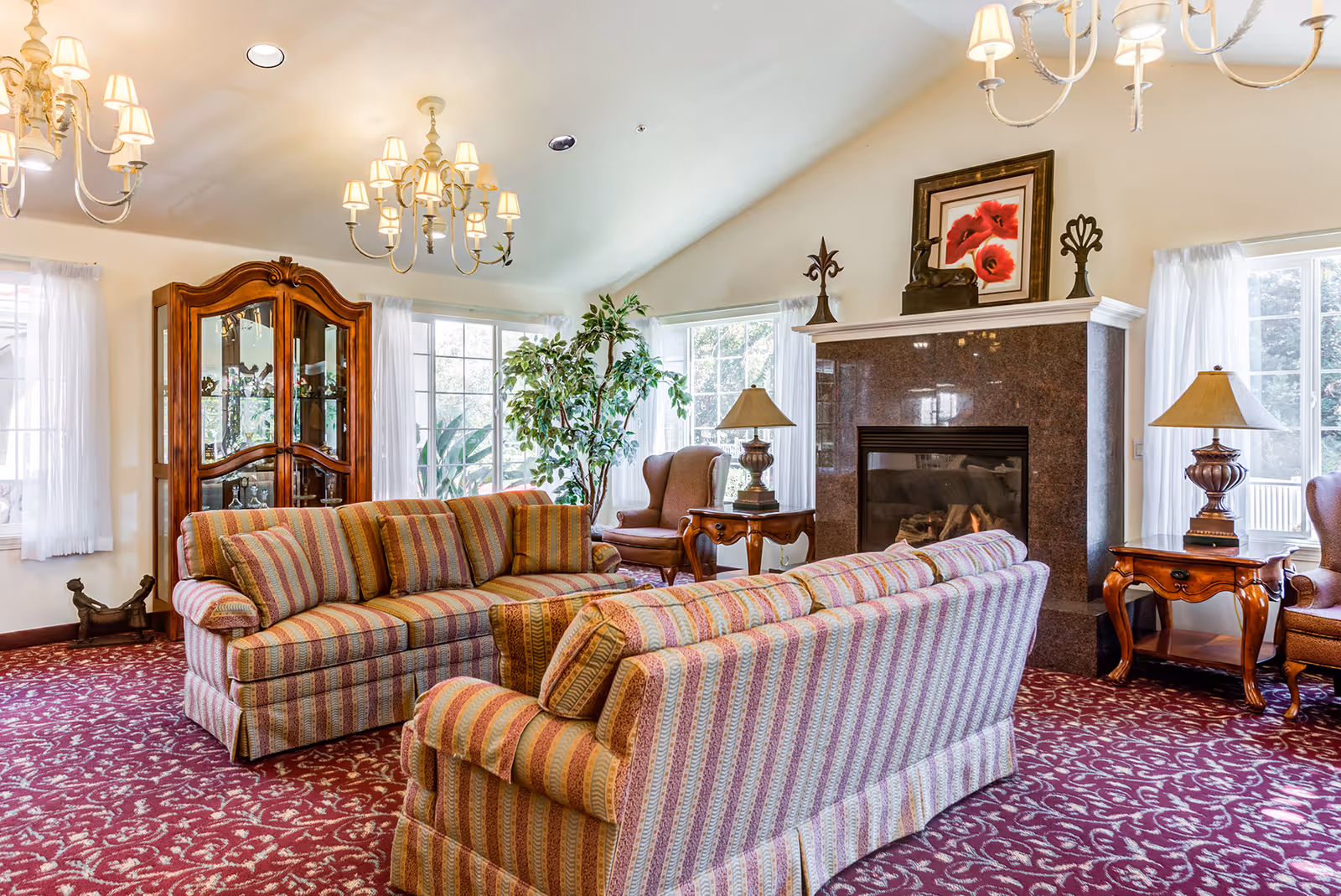 A cozy assisted living lounge with striped sofas arranged around a fireplace, chandeliers, side tables, a china cabinet, and patterned carpet.