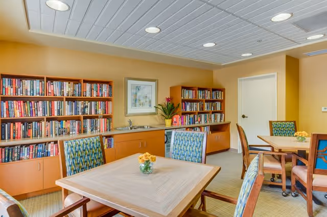 A cozy reading and activity room with wooden bookshelves filled with books, a small sink area with cabinets, and several tables with patterned chairs. The walls are painted a warm beige color, and there is a framed artwork and a potted plant on the countertop.