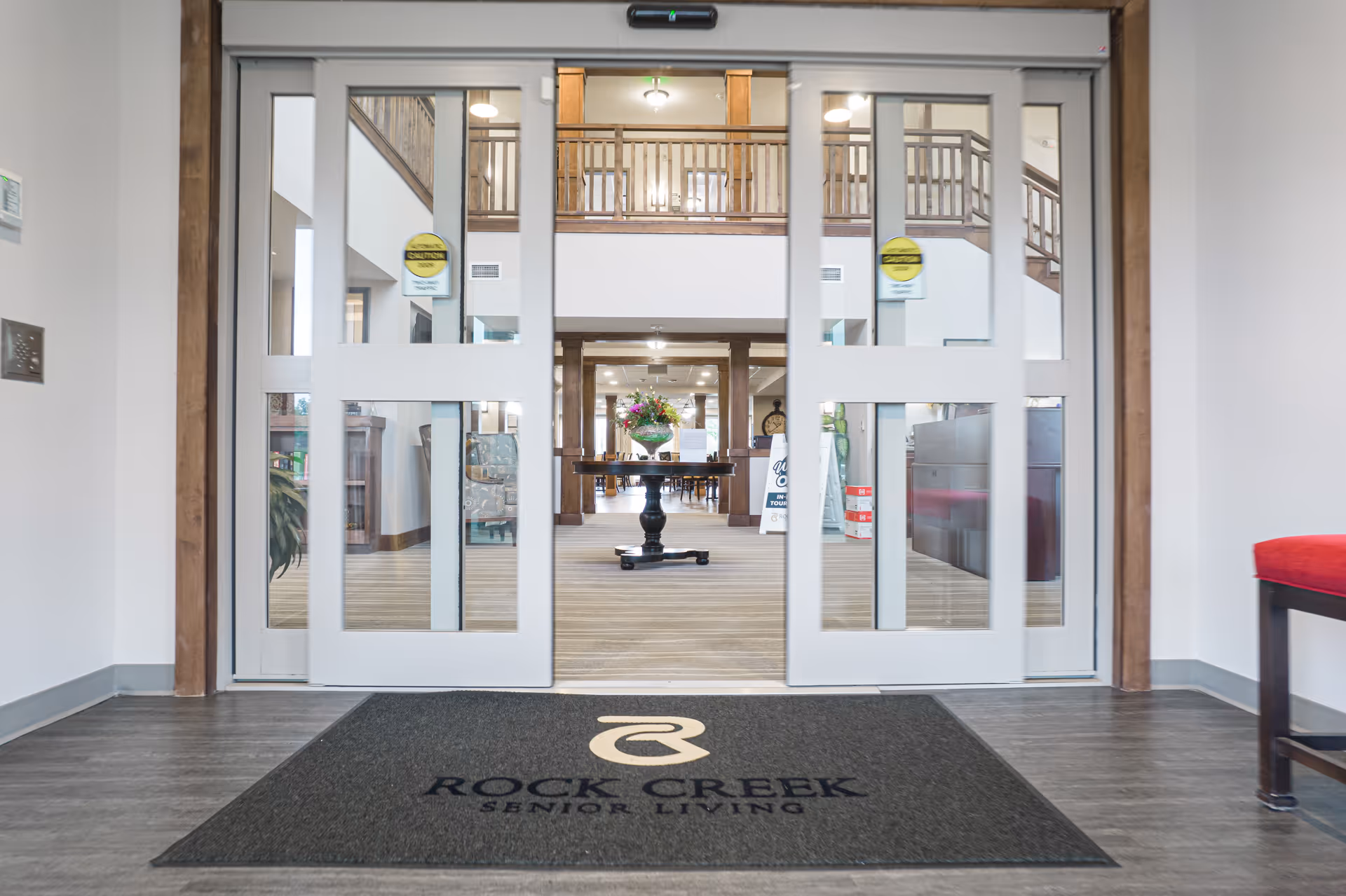 Entrance lobby with sliding glass doors opening to a reception area and a 'Rock Creek Senior Living' entry mat.