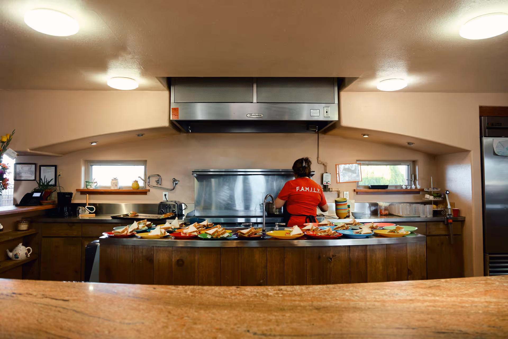 A kitchen area with a person wearing a red shirt labeled F.A.M.I.L.Y. standing at the sink. The countertop in front is filled with plates of sandwiches and food. The kitchen has wooden cabinets, two small windows, and stainless steel appliances including a large range hood and a refrigerator.