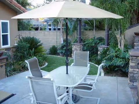 Outdoor patio with glass table, chairs, and umbrella among lush greenery.