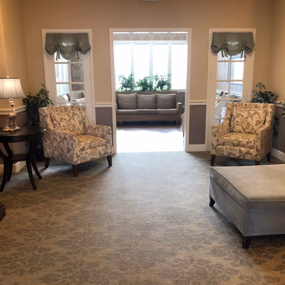 A cozy lounge with two patterned armchairs, an ottoman, a side table with lamp, and a sofa visible in a sunlit alcove.