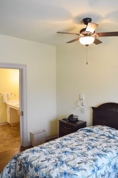 Small furnished bedroom with a single bed, nightstand and phone, ceiling fan, and an open door leading to a bathroom.