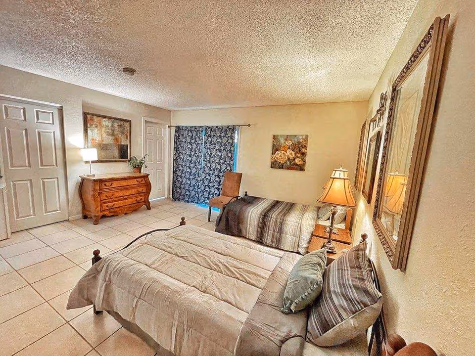 A bedroom with two beds, one with a beige comforter and the other with a striped comforter. There is a wooden dresser with a lamp and a plant on it, a chair near a window with blue patterned curtains, and framed artwork on the walls. The room has tiled flooring and a textured ceiling.