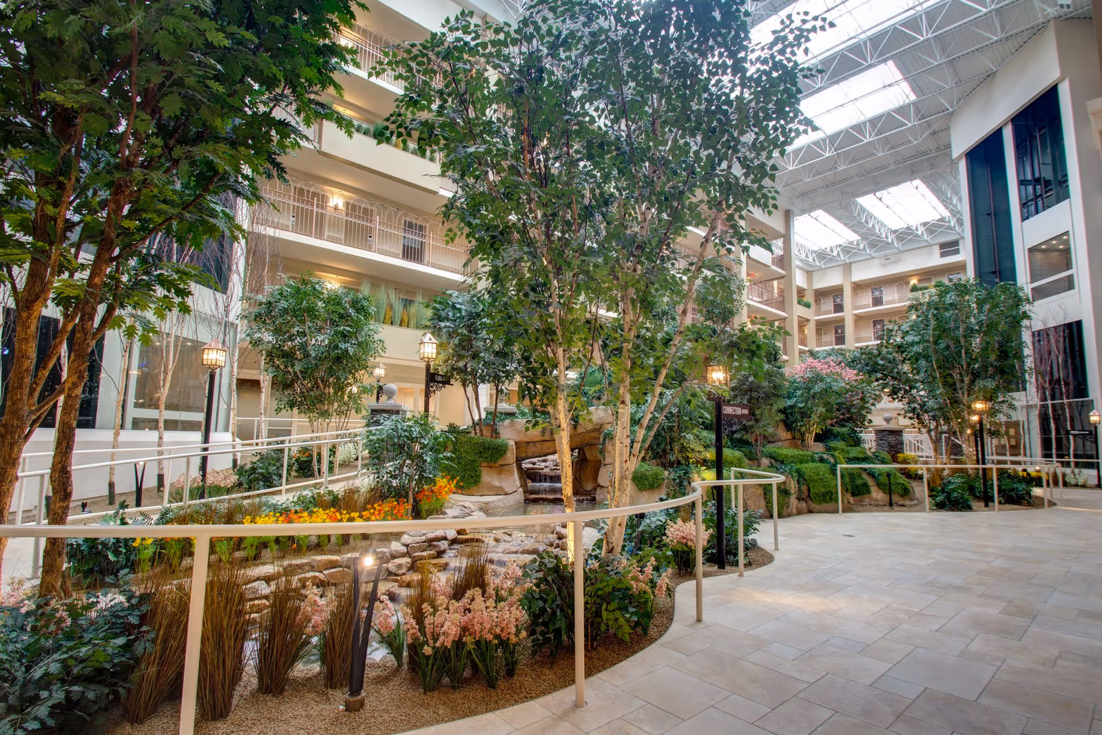 Indoor garden atrium in a senior living facility with lush green trees, flowering plants, a small waterfall feature, and a stone-lined stream. The space is surrounded by multiple floors with balconies and railings, and the ceiling has large skylights allowing natural light to fill the area.