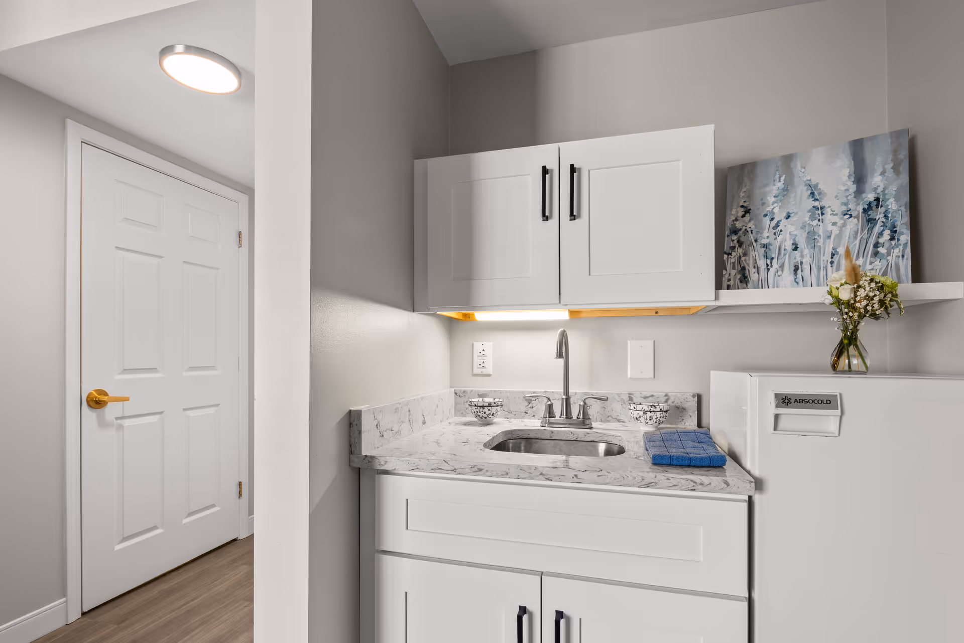 A small kitchenette area with white cabinets, a marble-patterned countertop, a stainless steel sink with a faucet, and a white mini refrigerator. There is a blue towel on the counter, a small vase with flowers, and a decorative painting on a shelf above the refrigerator. A white door with a gold handle is visible to the left.