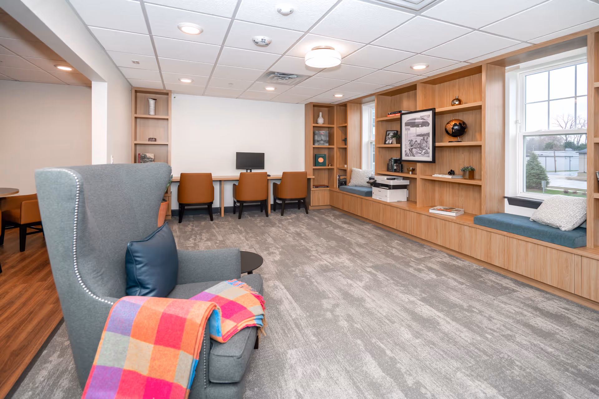 A bright and modern common area with a gray armchair draped with a colorful checkered blanket in the foreground. The room features built-in wooden shelves and window seats with cushions along the right wall. At the back, there is a long desk with three brown chairs and a computer monitor. The floor is carpeted in a light gray pattern, and the ceiling has recessed lighting.