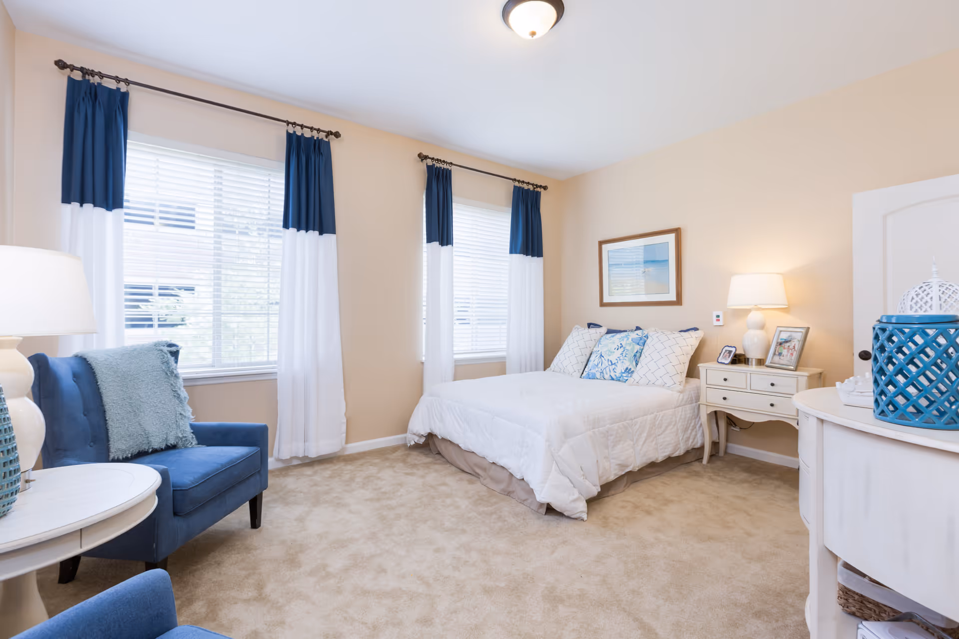 A bright and cozy bedroom with beige walls and carpet. The room features a bed with white bedding and decorative pillows, two windows with white and navy blue curtains, a blue armchair with a light blue throw pillow, a white nightstand with a lamp and framed photos, and a white dresser with blue decorative items.
