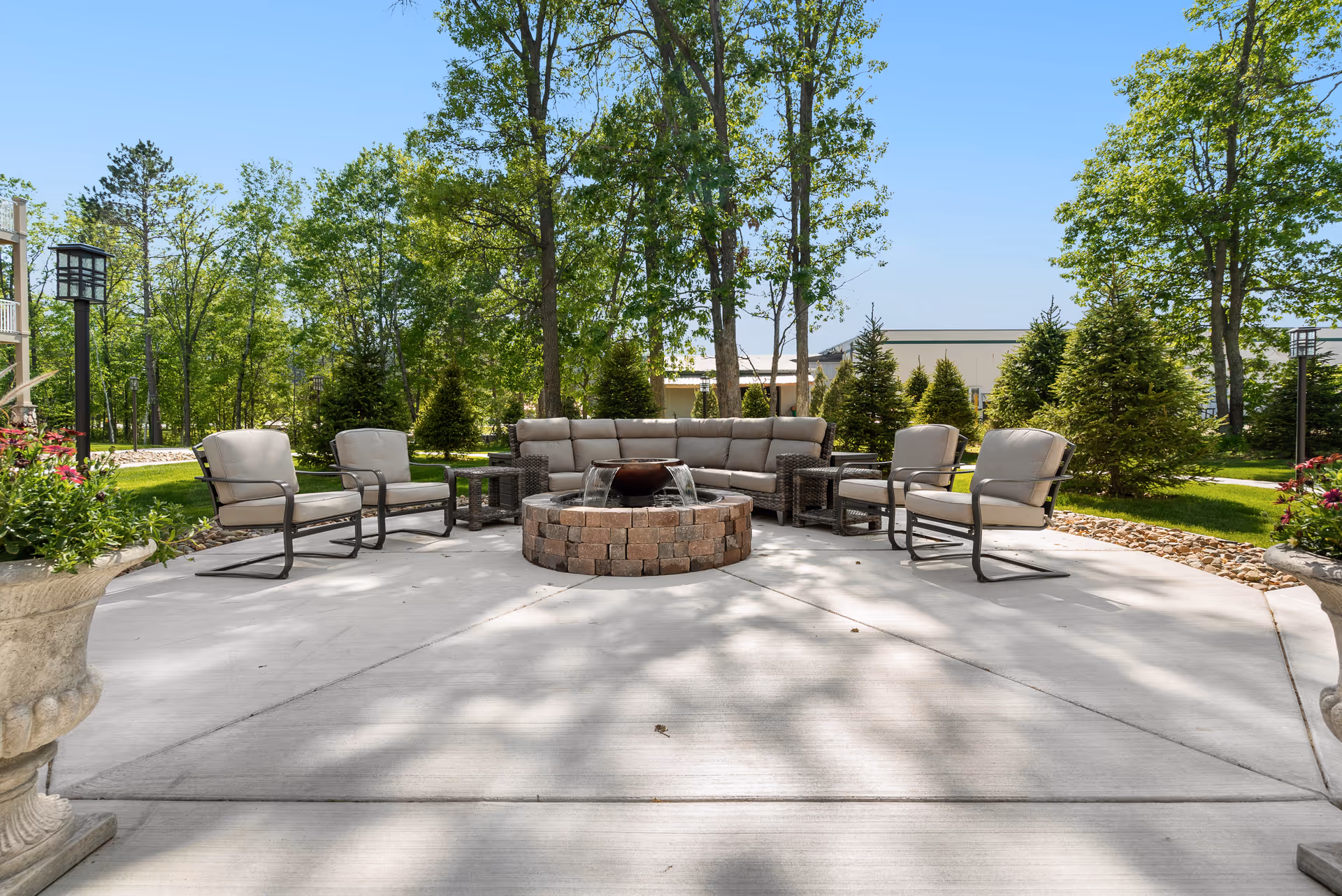 Outdoor patio with a circular brick fire pit surrounded by cushioned chairs and a sofa amid trees and landscaped greenery.