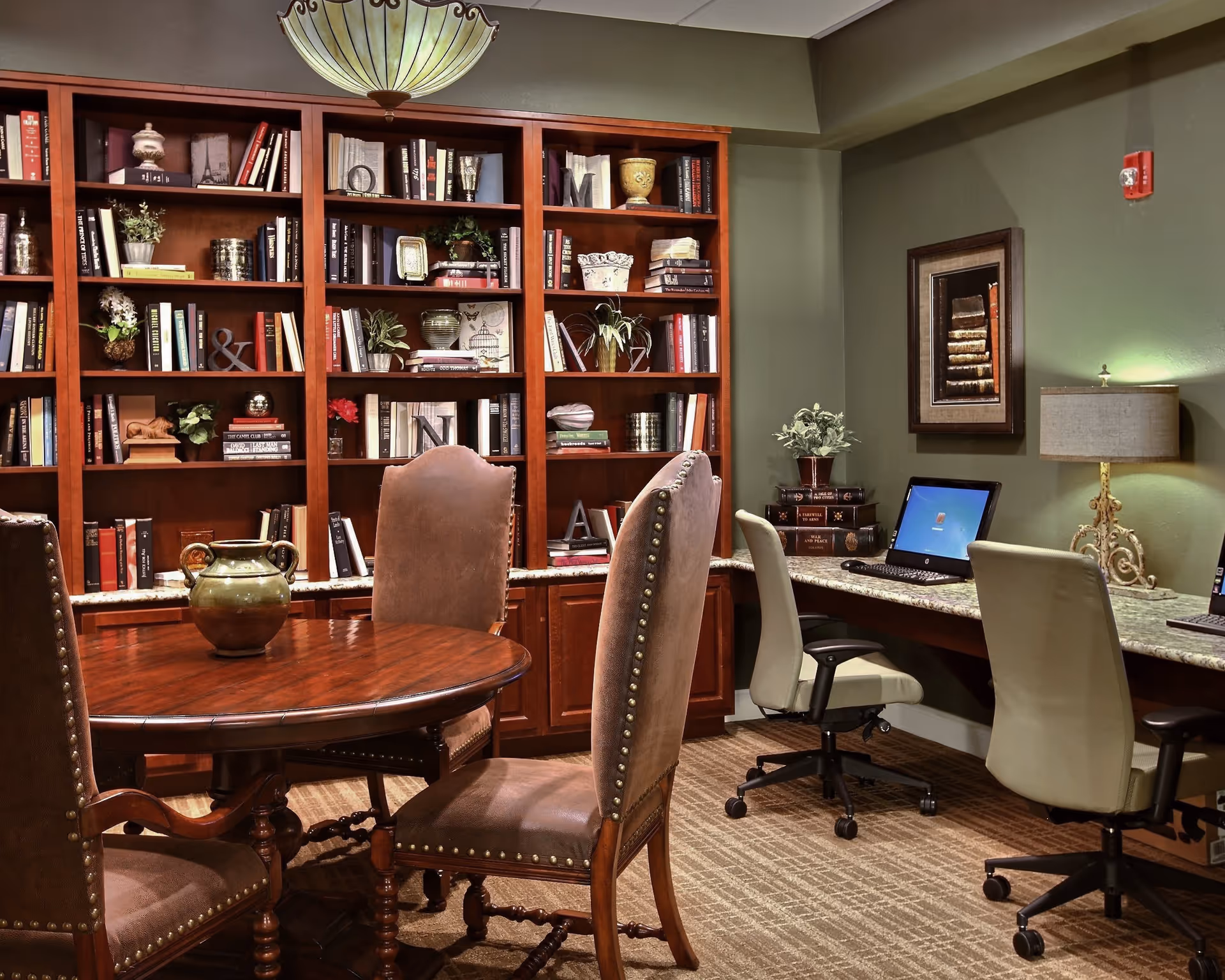 A cozy room with a round wooden table surrounded by four upholstered chairs in the foreground. Behind the table is a large wooden bookshelf filled with books and decorative items. To the right, there is a long desk with two office chairs and two desktop computers. The walls are painted green, and there is a framed picture and a table lamp on the desk.