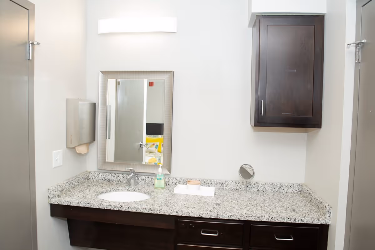 A bathroom vanity area with a granite countertop, an undermount sink, a chrome faucet, a rectangular mirror with a silver frame, a wall-mounted dark wood cabinet, a soap dispenser, a small round mirror, and a paper towel dispenser on the left wall.