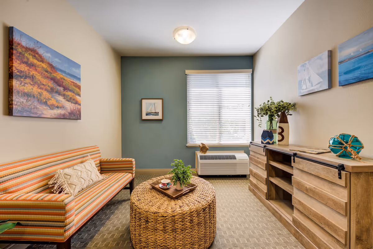 A cozy living room with a striped orange and beige sofa, a round woven coffee table with a small plant and a cup on a tray, a wooden cabinet with decorative items including vases and artwork on the walls. A window with blinds lets in natural light, and the walls are painted beige and teal.