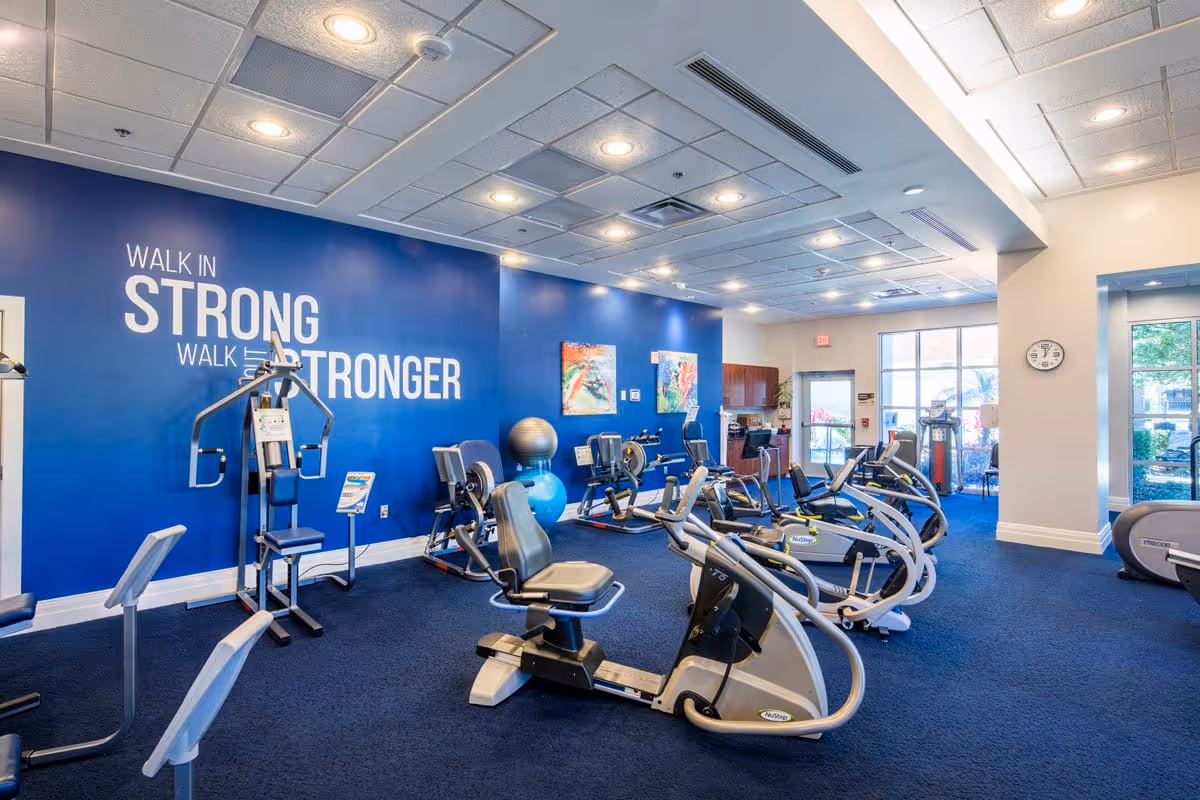 A fitness room with various exercise equipment including recumbent bikes and strength machines. The walls are painted blue and white, with motivational text on the blue wall that reads 'WALK IN STRONG WALK OUT STRONGER'. There are two colorful abstract paintings on the blue wall, exercise balls, and large windows letting in natural light.