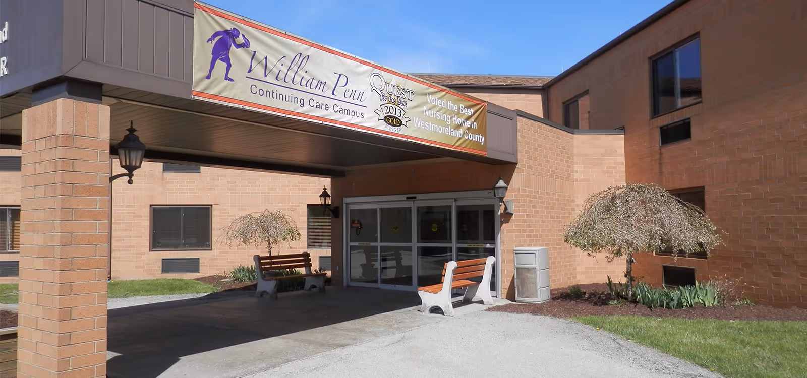 Entrance of William Penn Continuing Care Campus with a covered drop-off area, two benches, a small tree, and a banner above the entrance that reads 'William Penn Continuing Care Campus' and mentions an award for the best nursing home in Westmoreland County.