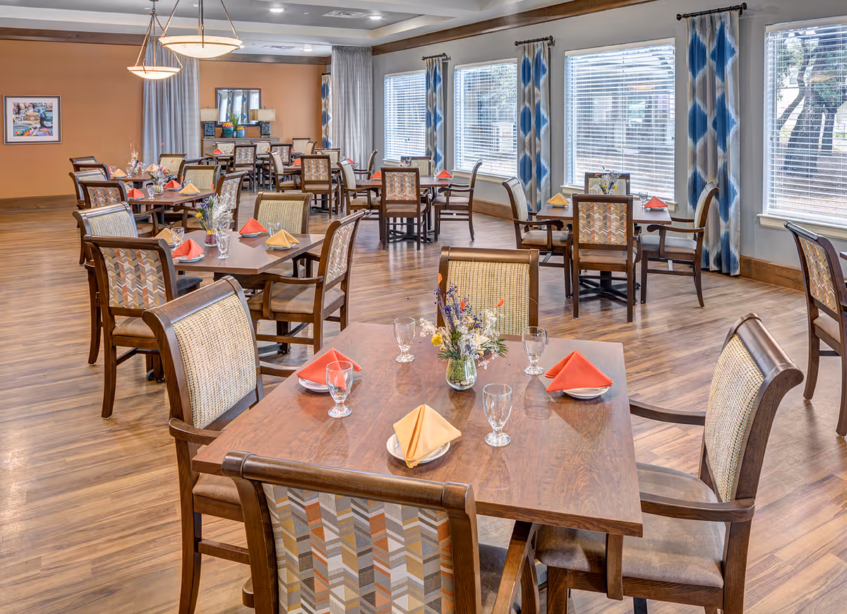 A bright and spacious dining room with multiple wooden tables and cushioned chairs. Each table is set with folded napkins in red and yellow, glassware, and small floral centerpieces. Large windows with blue and beige patterned curtains allow natural light to fill the room. The floor is wooden, and the walls are painted in warm tones with some artwork and mirrors.
