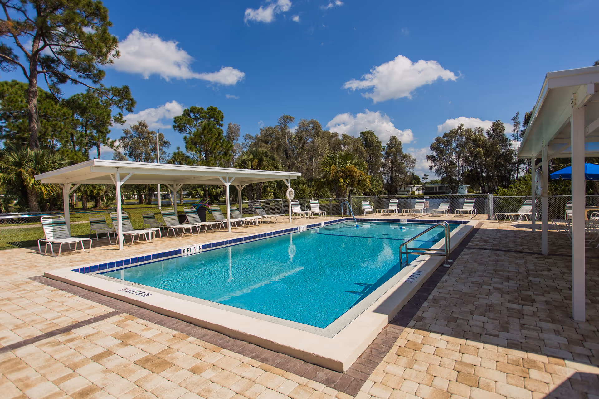 Outdoor swimming pool with clear blue water surrounded by a paved deck. Several lounge chairs are arranged under a white canopy and around the pool area. Trees and greenery are visible in the background under a partly cloudy blue sky.