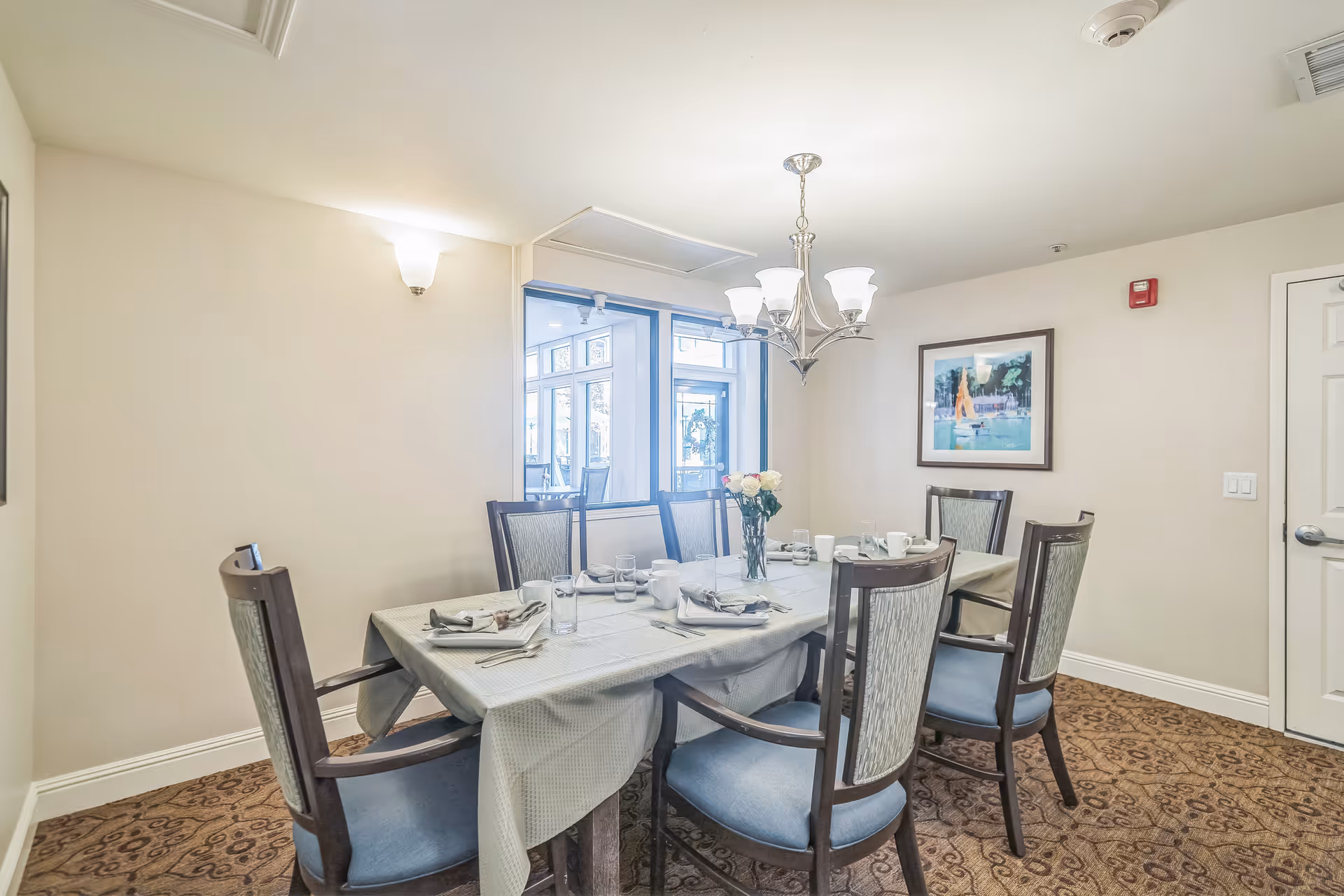 A dining room with a rectangular table covered with a light-colored tablecloth, set with plates, glasses, and napkins. Six wooden chairs with blue upholstered seats surround the table. A chandelier with five lights hangs above the table. There is a framed picture on the wall and a door to the right. A window shows another room with chairs and a door with a wreath.