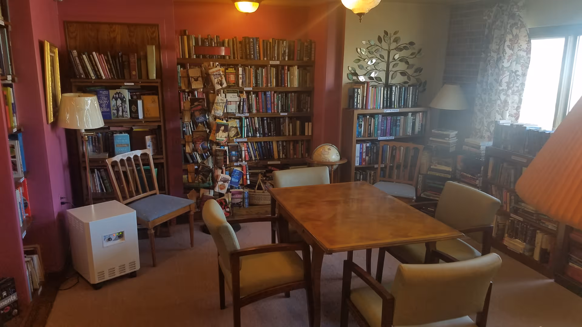 A cozy room with wooden bookshelves filled with books, a wooden table surrounded by six chairs, two floor lamps, a globe on a small table, and a window with floral curtains letting in natural light.