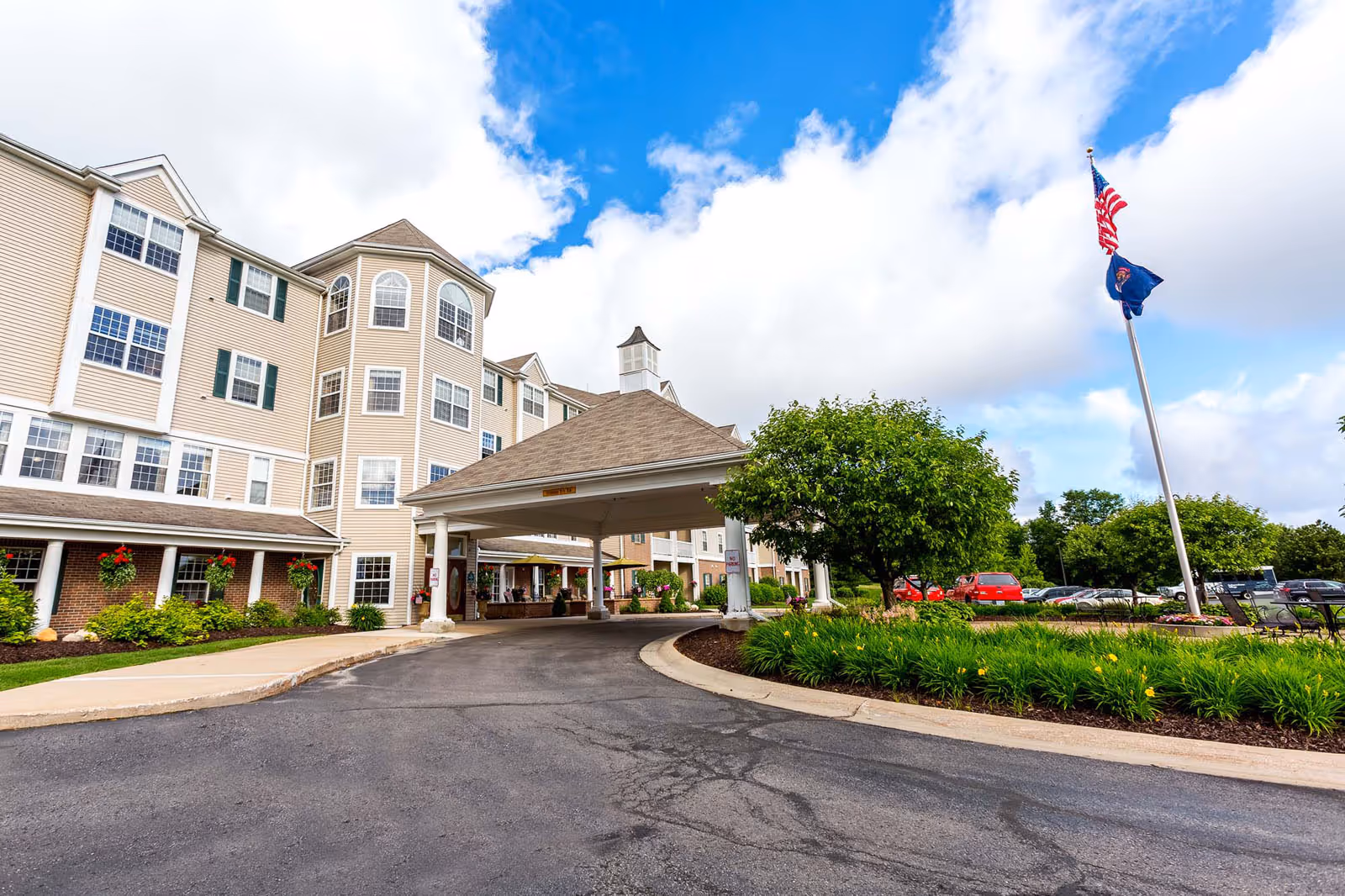 Multi-story senior living building with a covered porte-cochère entrance, landscaped driveway, and flagpoles under a partly cloudy sky.
