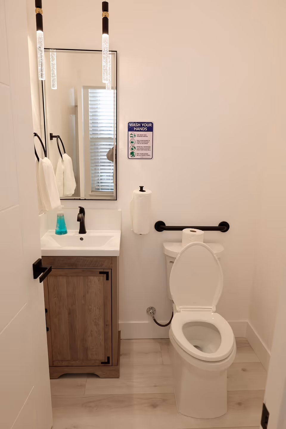 Small accessible bathroom with a sink and mirror on the left, a toilet with a grab bar on the right, hand soap, paper towel holder, and a 'Wash Your Hands' sign on the wall.