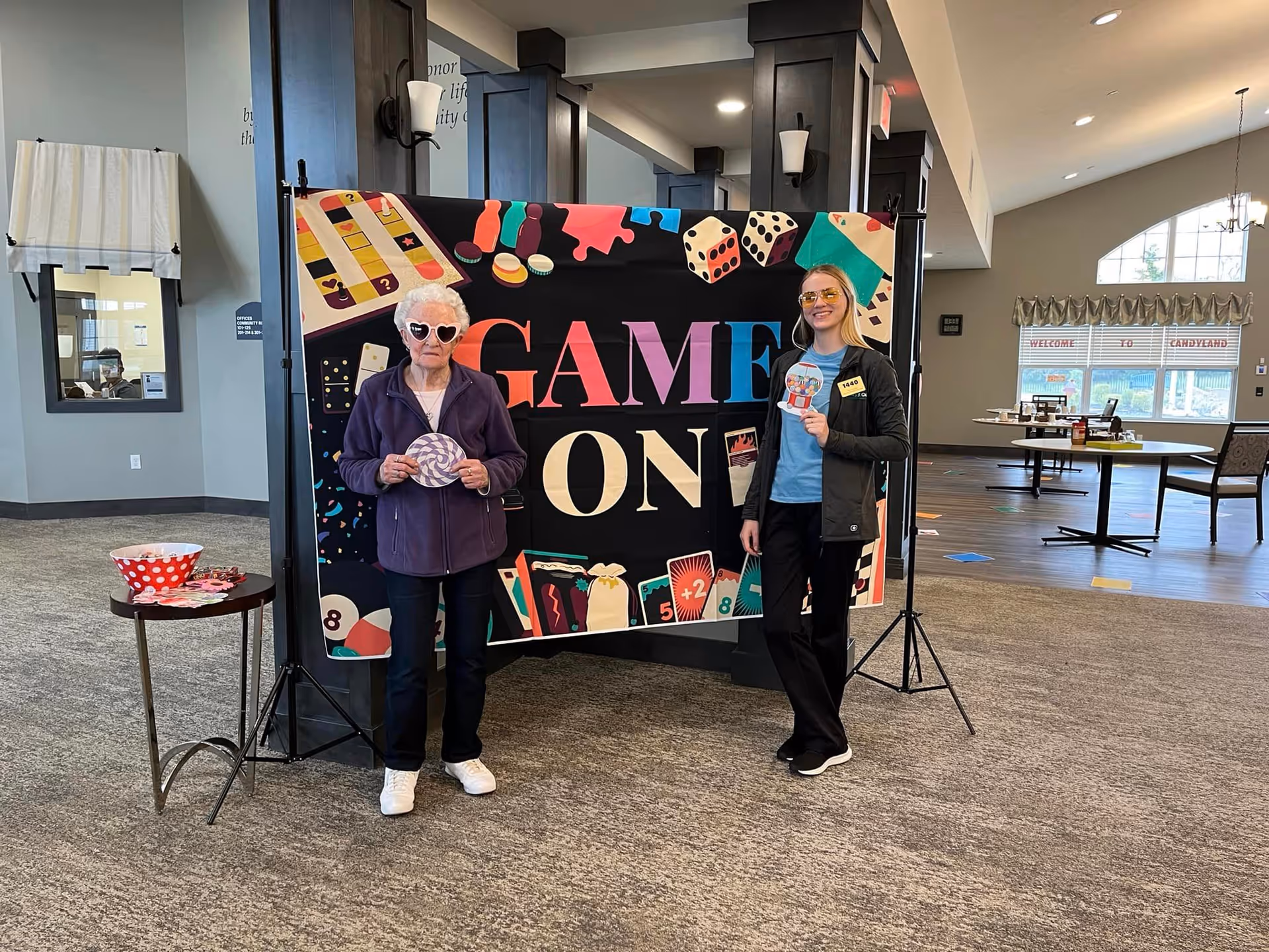 Two women standing in front of a colorful banner that reads 'GAME ON' with various board game illustrations. One woman is elderly with white hair and heart-shaped sunglasses, wearing a purple jacket and holding a circular game piece. The other woman is younger, wearing yellow-tinted glasses, a black jacket, and holding a card. They are in a spacious room with tables and chairs in the background.