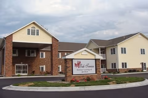 Exterior view of Oak Terrace Senior Living of Jordan building with a brick and beige siding facade, a covered entrance, and a sign in front displaying the facility name surrounded by a landscaped area with grass and flowers under a cloudy sky.