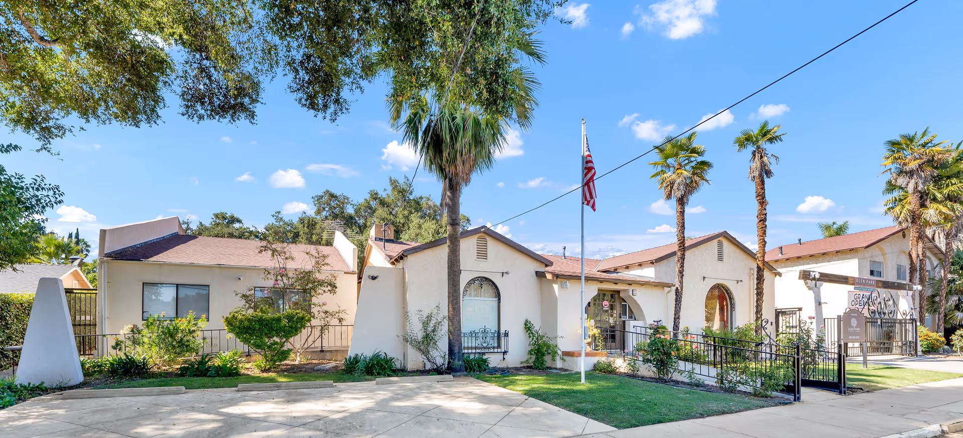 Front exterior of a single-story senior living building with arched entrances, palm trees, a flagpole, and a gated walkway.