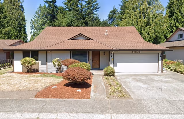 Front exterior view of a single-story house with a brown roof, white siding, a wooden front door, a two-car garage, and a small landscaped yard with bushes and trees.