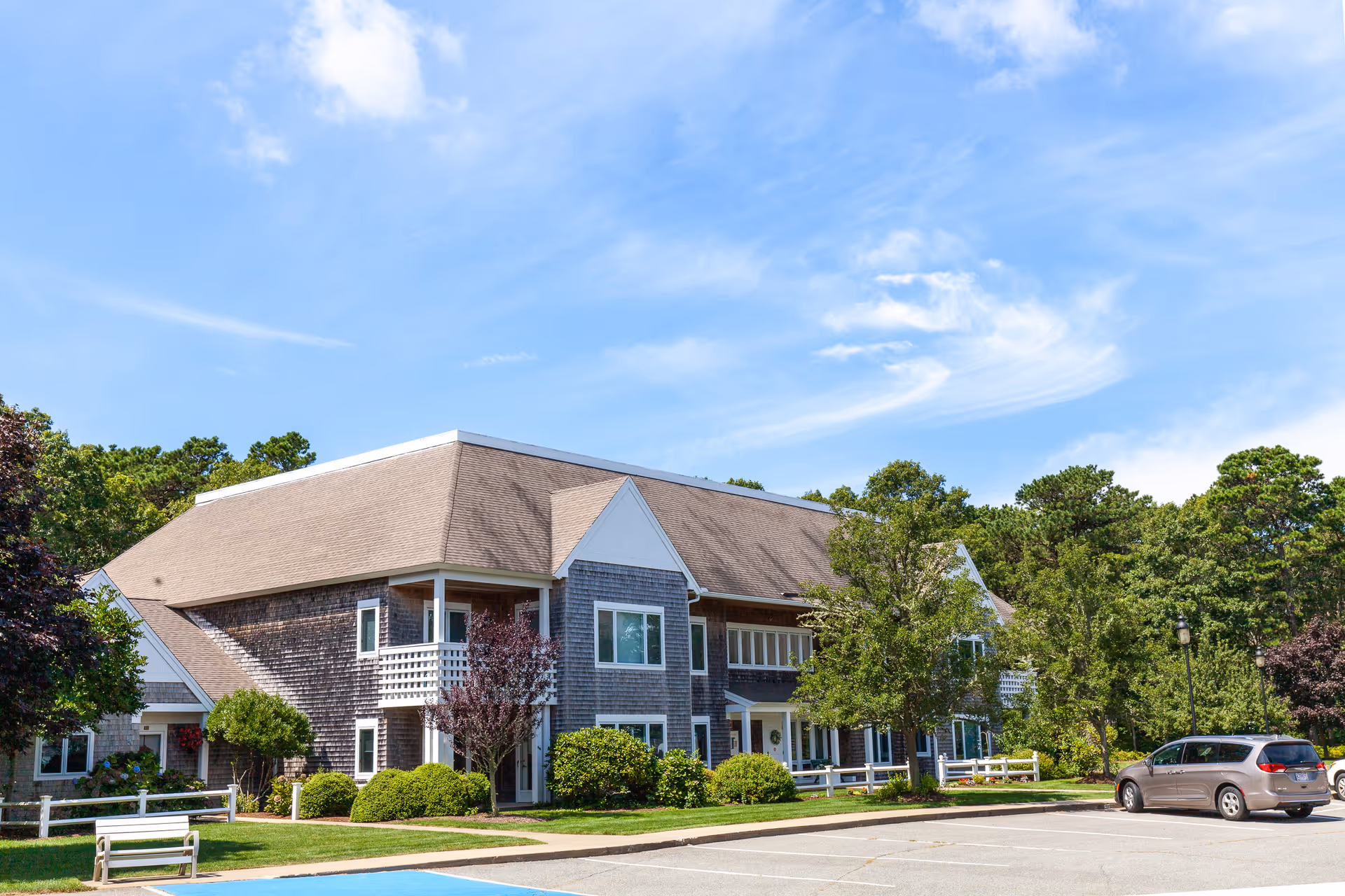 Exterior view of a two-story senior living facility building with a sloped roof, surrounded by trees and shrubs, with a parking lot in front under a partly cloudy blue sky.
