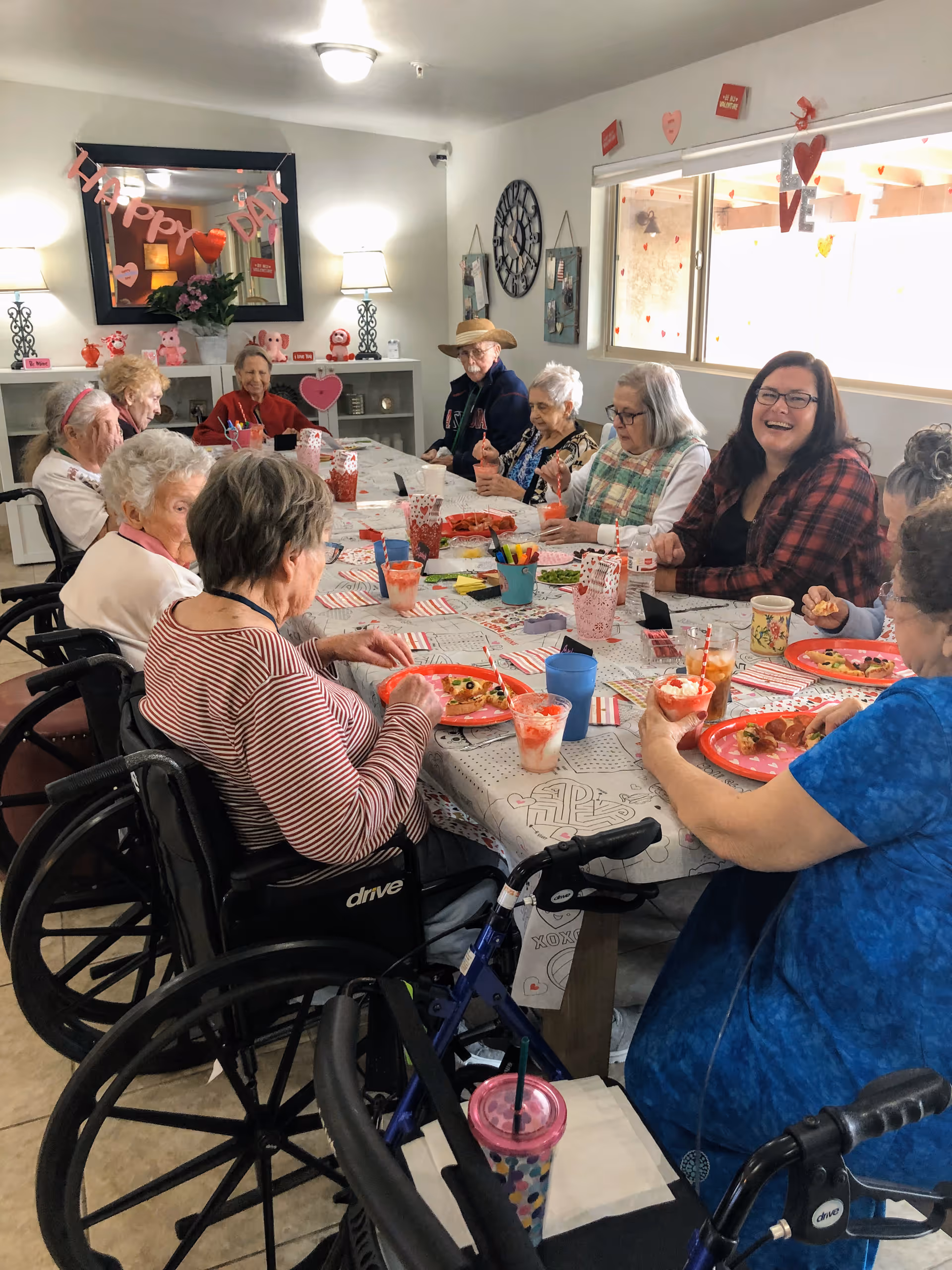 A group of elderly people and a caregiver sitting around a long table in a decorated room, enjoying a meal together. The table is covered with a festive tablecloth and red plates with food. The room has Valentine-themed decorations including hearts and banners that say 'Happy Day' and 'LOVE'. Some of the elderly individuals are in wheelchairs.