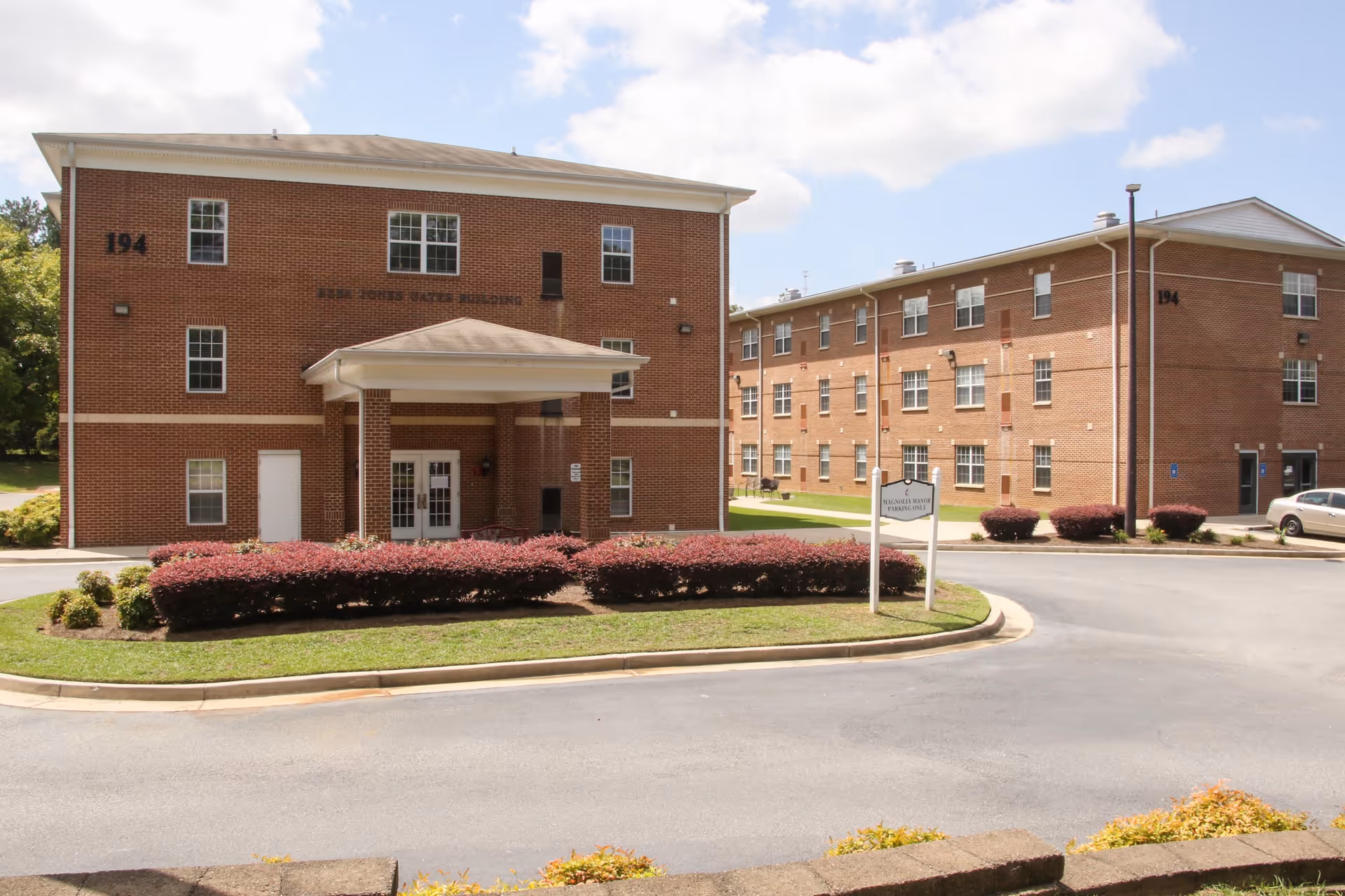 Exterior view of two three-story brick buildings with multiple windows, surrounded by a paved driveway and landscaped bushes. A sign in front reads 'Magnolia Manor Parking Only'. The sky is partly cloudy.