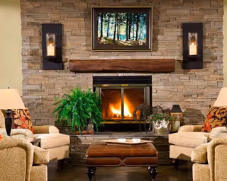 Cozy living room featuring a stone fireplace with a lit fire, armchairs, an ottoman, plants, and wall sconces.