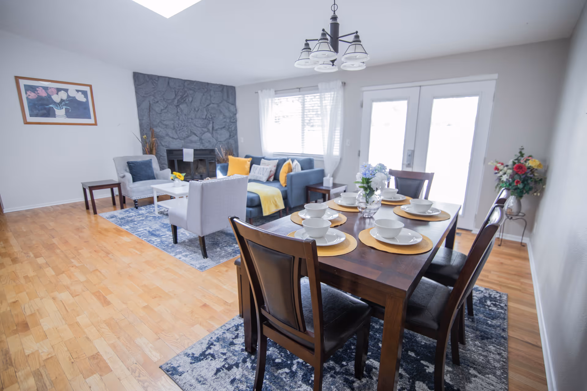 Bright open living and dining area with a set wooden dining table in the foreground and a seating area with sofa and fireplace in the background.