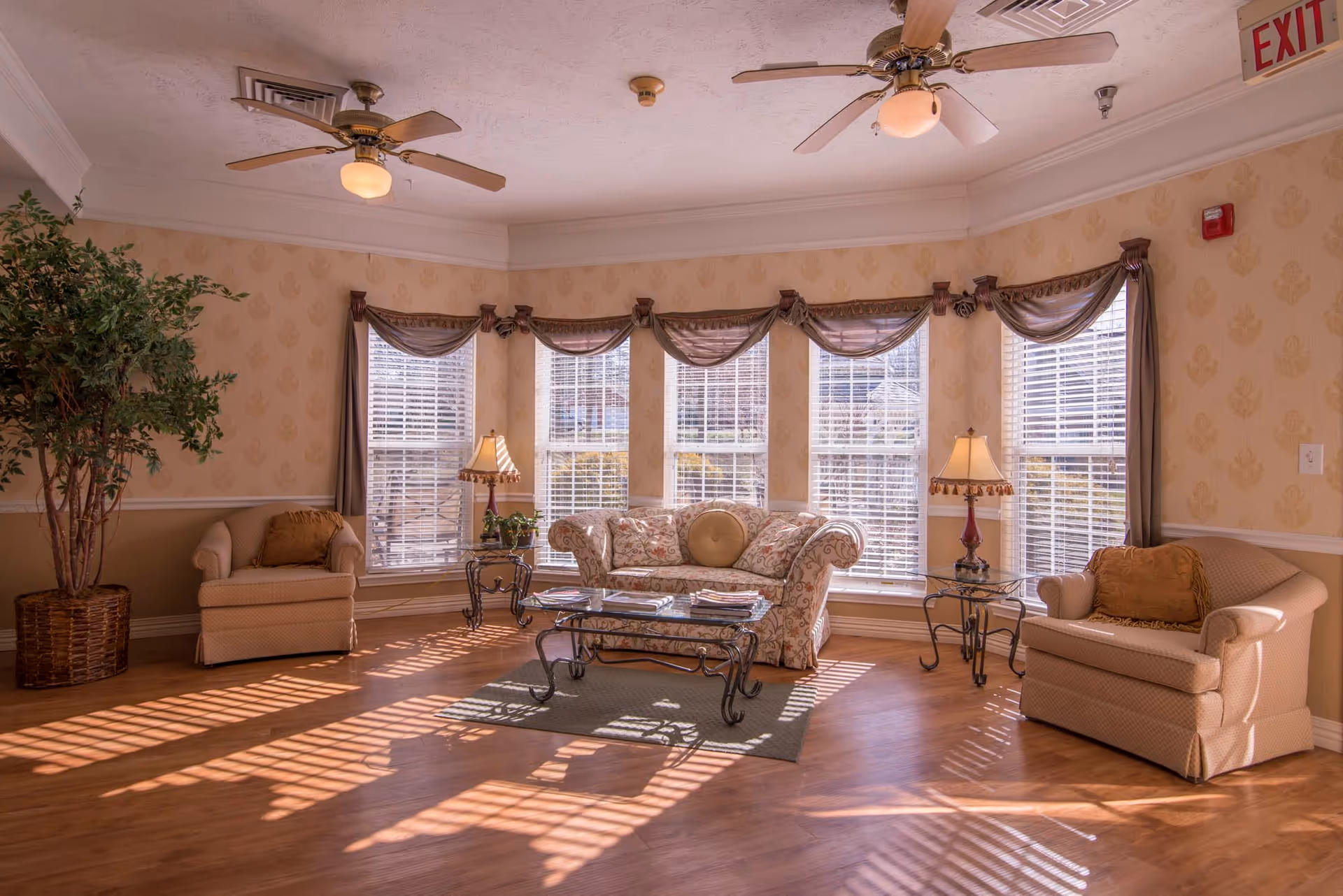 A cozy living room area with a floral-patterned sofa, two beige armchairs, two side tables with lamps, a glass coffee table on a small rug, large windows with blinds and draped curtains letting in sunlight, two ceiling fans, and a large potted plant in the corner.