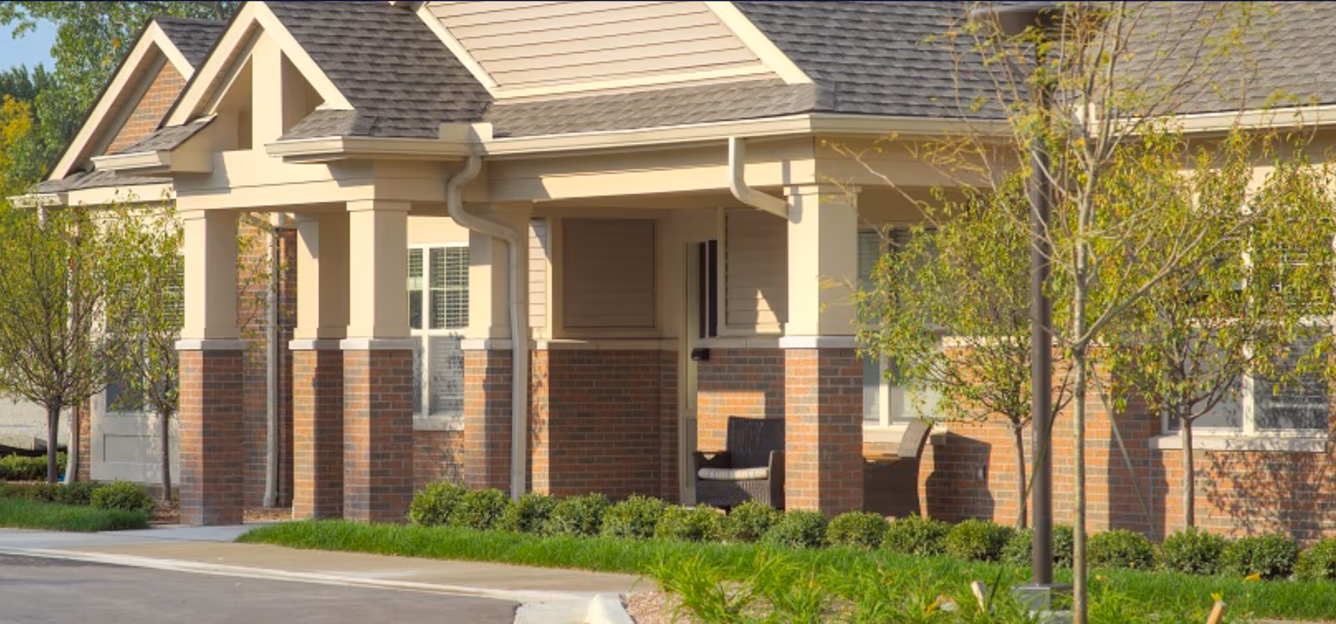 Exterior view of a senior living facility building with brick and beige siding, a covered porch with seating, small trees, and well-maintained landscaping.