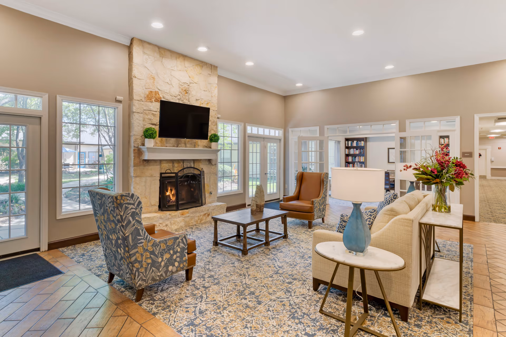 Spacious senior living lounge with sofas and armchairs arranged around a stone fireplace and wall-mounted TV.