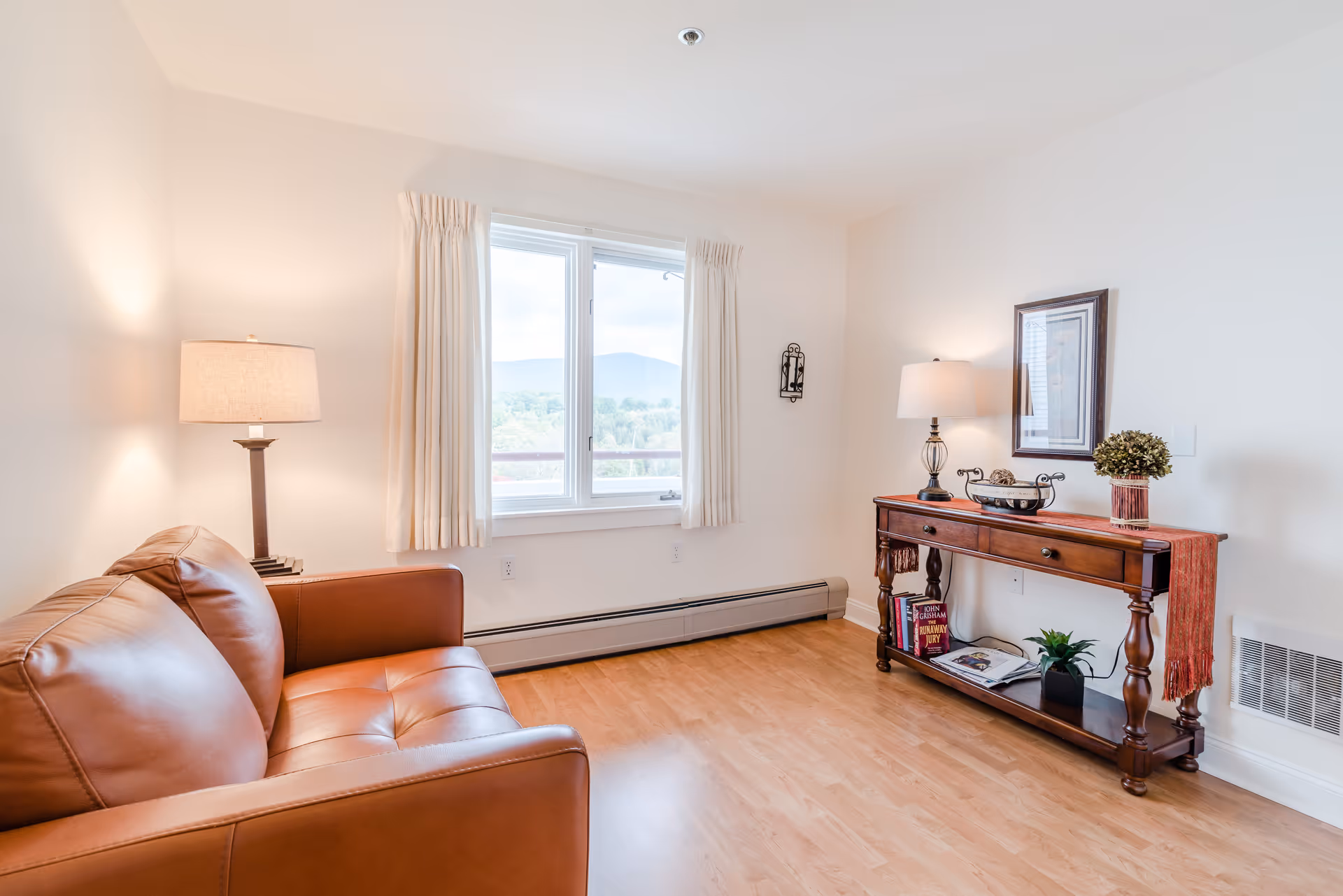 Bright living room with a leather sofa, a window with curtains, two table lamps, and a wooden console table with decor.