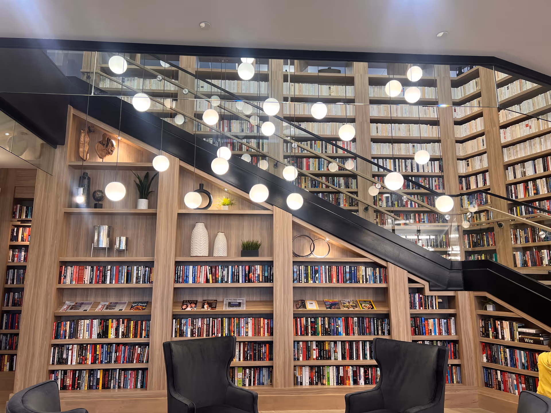 Interior view of a library or reading area with tall wooden bookshelves filled with books extending up to the ceiling. A black staircase with glass railing runs diagonally across the shelves. Several round pendant lights hang from the ceiling, reflecting in the glass. Two black armchairs are positioned in front of the bookshelves.