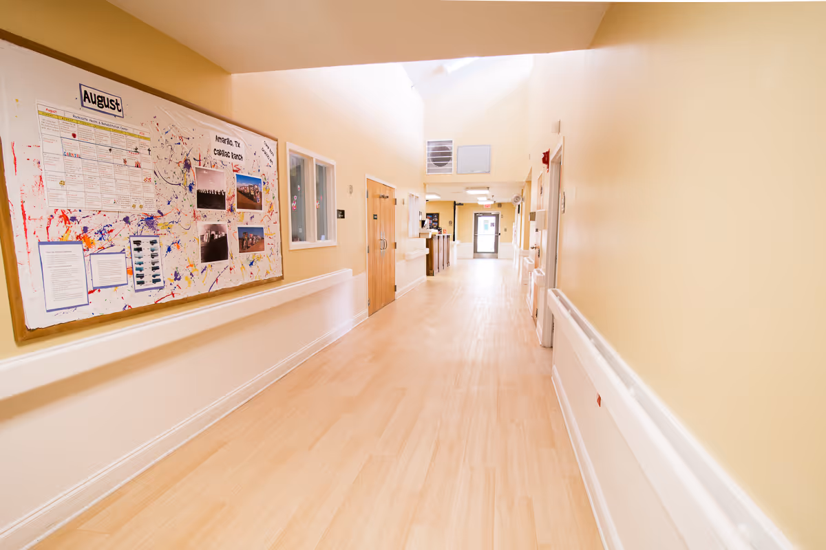 A bright, clean hallway in a senior living facility with light wood flooring and pale yellow walls. On the left wall, there is a large bulletin board with various notices and colorful decorations. Several doors and windows line the hallway, and at the far end, there is a door with a window letting in natural light.