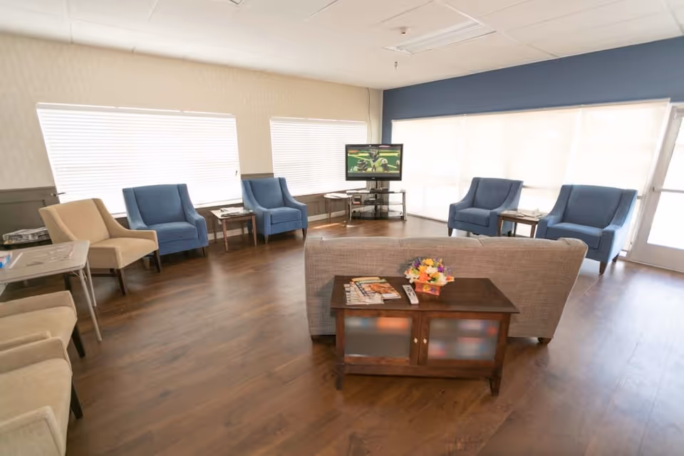 A bright and spacious living room with wooden flooring, featuring a gray sofa facing a TV on a stand. Surrounding the sofa are several armchairs in blue and beige, with small side tables placed between them. Large windows with blinds allow natural light to fill the room.