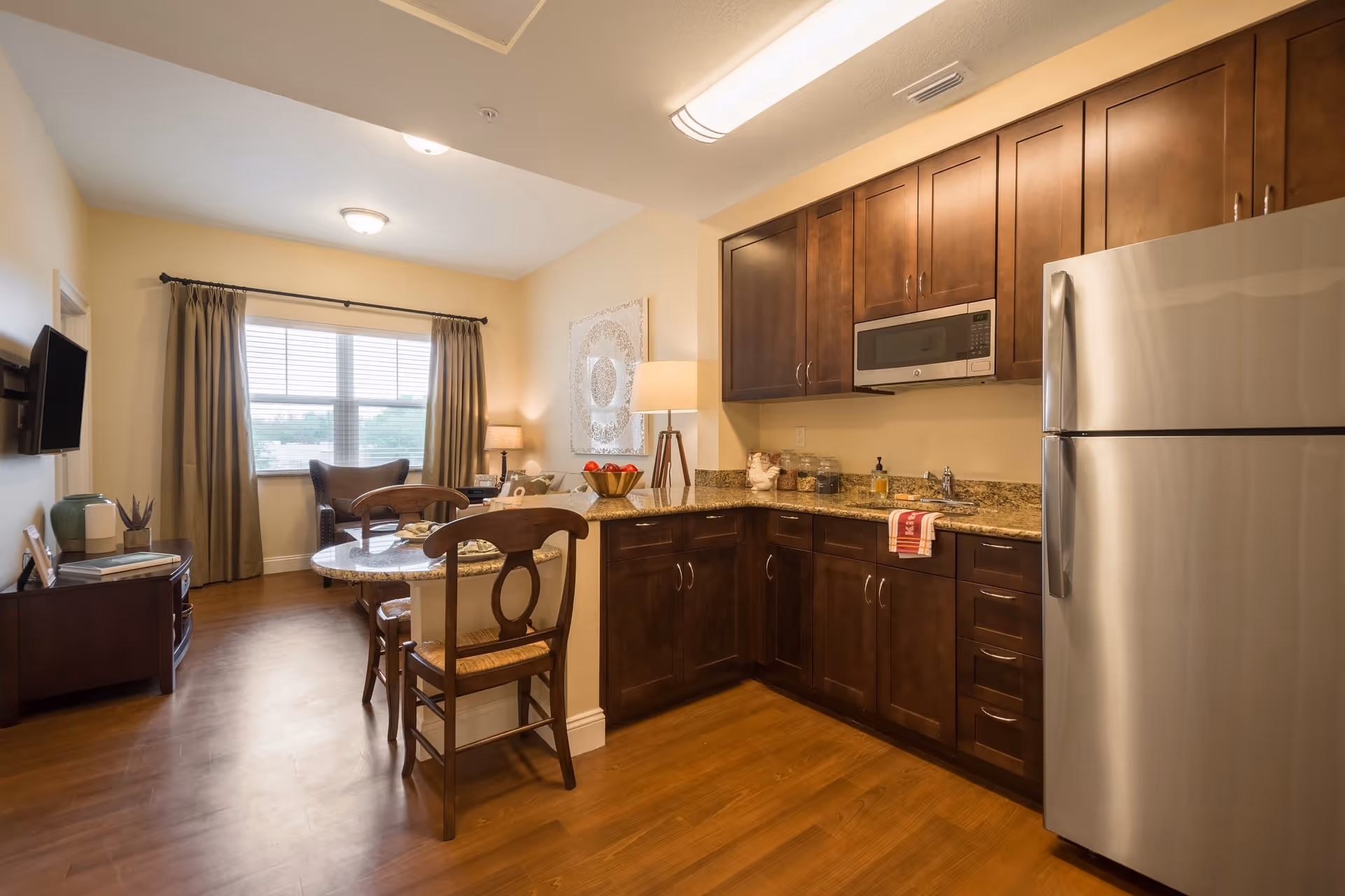 A cozy kitchen and living area in a senior living facility featuring wooden cabinets, a stainless steel refrigerator, a microwave, a granite countertop with two wooden chairs, and a small living space with a window, armchair, TV, and side table with lamps.