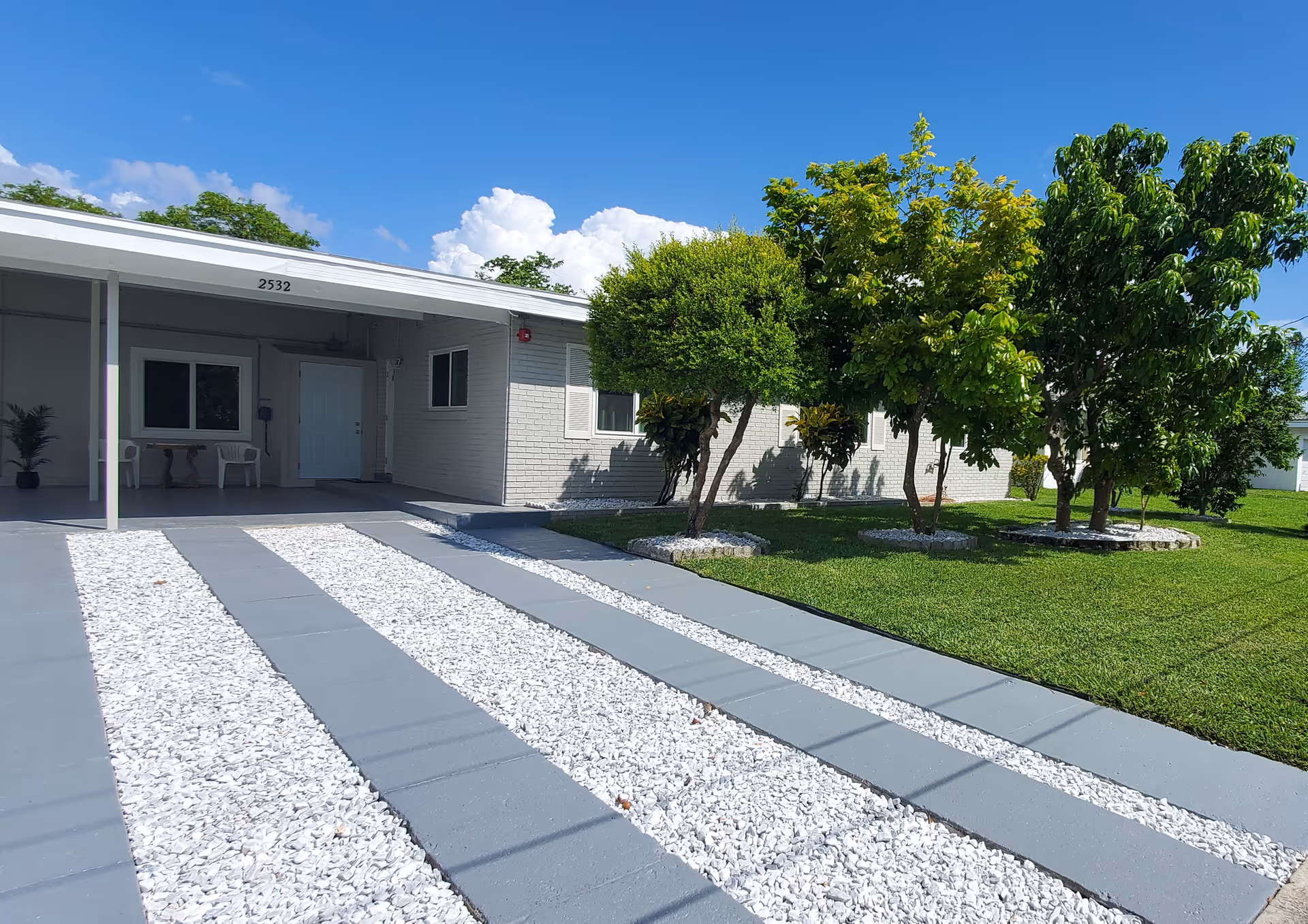 Front exterior view of a single-story building with a covered entrance, white door, and windows. The driveway has gray concrete strips with white gravel in between. There are several trees and a well-maintained green lawn on the right side under a clear blue sky.