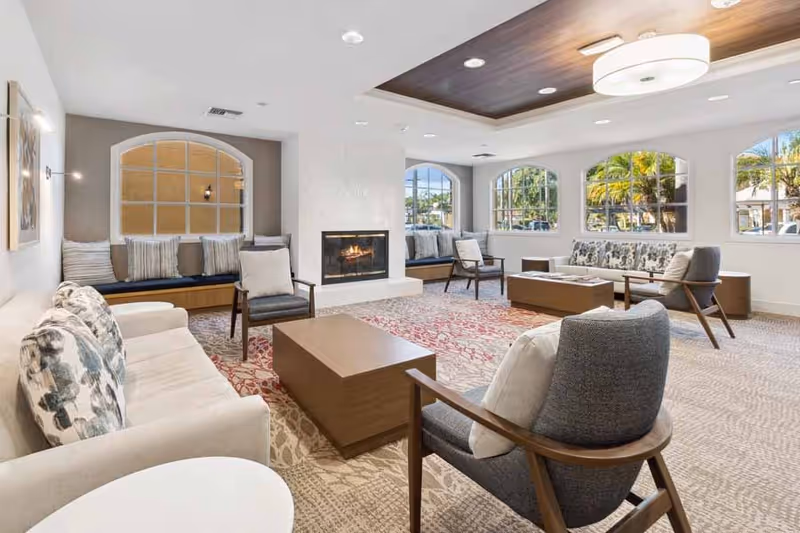 A bright and spacious living room area with multiple seating options including sofas, armchairs, and built-in window benches with cushions. The room features a modern fireplace in the center, large windows letting in natural light, a patterned carpet, and a wooden ceiling accent with recessed lighting.