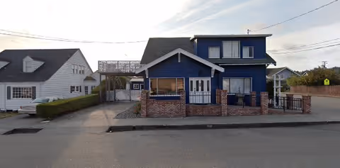 Exterior view of a two-story blue house with a brick fence in front, located on a street with neighboring houses visible on either side under a partly cloudy sky.